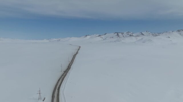 Drone view of a long empty road cutting through the snow-covered Arabel Plateau, Kyrgyzstan, with distant mountain ridge and overcast winter sky