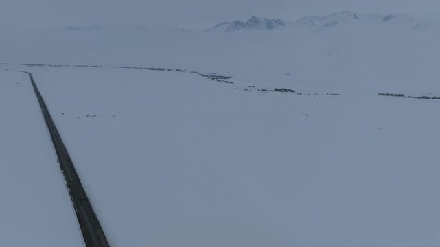 Drone footage of a long empty highway cutting across a vast snow-covered plateau at Ala-Bel Pass, Jalal-Abad Region, Kyrgyzstan, with distant mountains and overcast sky