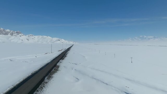 Drone view of a long asphalt road cutting through the snow-covered Alay Valley, with distant mountain ridges under a clear blue sky in Osh Region, Kyrgyzstan