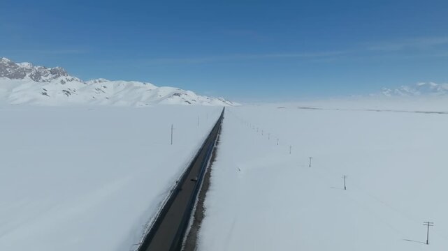Drone footage of a long straight highway cutting through deep snow in the Alay Valley, Osh Region, Kyrgyzstan, with distant peaks under a clear blue sky
