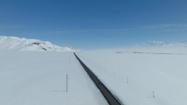 Drone footage of a straight highway cutting through vast snowy plains with distant mountains under clear blue sky in Alay Valley, Osh Region, Kyrgyzstan