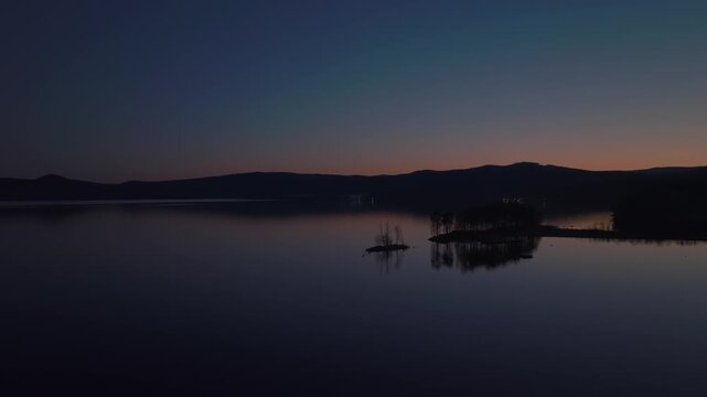 Aerial footage of Lake Turgoyak at dusk with a small wooded island, smooth reflective water and dark mountain silhouette, peaceful natural landscape in Chelyabinsk Oblast, Russia