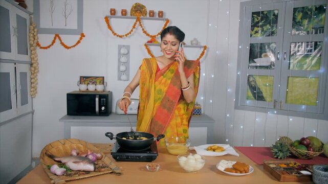 A newly wedded girl busy talking with her mother on mobile while cooking. A medium shot of a traditional Bengali housewife frying cutlets in her kitchen and talking on her smartphone