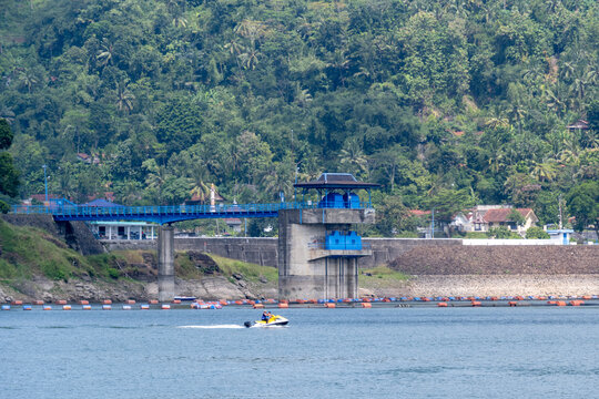 A water intake tower and concrete bridge at Sempor Reservoir, with a person riding a jet ski across the calm waters against a lush green hillside backdrop, Kebumen, Central Java, Indonesia.