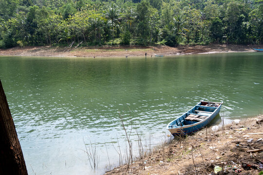A small blue motorized wooden boat moored on the grassy shore of Sempor Reservoir, with calm open water and lush green hills in the background, Kebumen, Central Java, Indonesia.