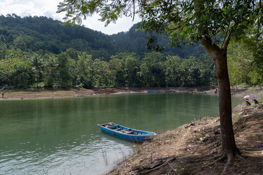 A small blue wooden boat moored on the grassy shore of Sempor Reservoir, framed by tree branches with calm green water and lush tropical hills in the background, Kebumen, Central Java, Indonesia.
