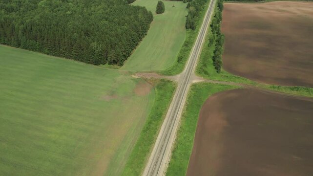 Drone footage looking down on a quiet country road cutting through farmland, green meadows and dense forest, showing summer landscape patterns and rural transport