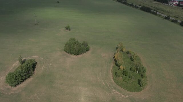 Drone footage of wide green meadow and farmland by a calm lake, with a rural road, power lines, and forest edge in the distance on an overcast summer day