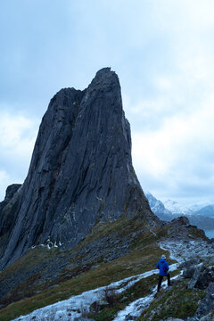 A lone hiker ascends the snowy, rugged path of the Segla trail towards the dramatic gray rock peak under heavy clouds in Norway.