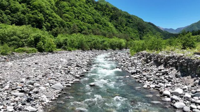 Clear stream rushes over smooth stones. Lush green hills rise behind the water. Sky is bright blue, cloudless above. Riverside feels peaceful, untouched. Perfect for tranquil nature scenes
