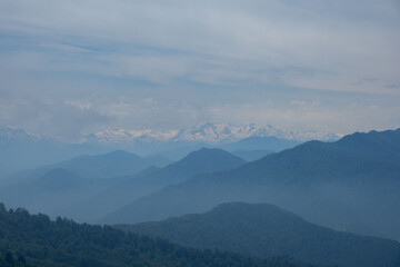 Fototapeta premium Distant snow-capped Caucasus mountain peaks behind misty blue layers of hills under a cloudy sky