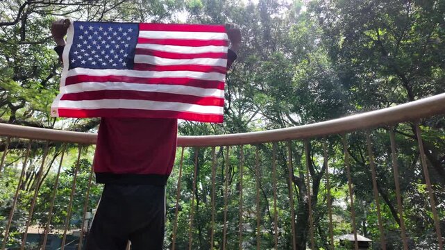 Close up tracking shot focusing on the red and white stripes of the American flag waving gently as it hangs from a man's shoulders while standing on an outdoor balcony surrounded by green trees.
