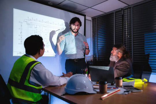 Engineering team discussing construction plan in dark meeting room, Construction manager explaining infrastructure plan with senior manager brainstorming over house blueprint projection in office.