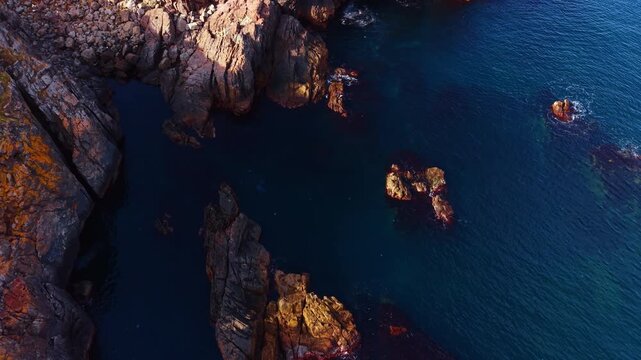 Rugged sea rocks and narrow inlet viewed from above. Drone perspective of weathered orange and grey boulders, dark tidal pool and turquoise water.