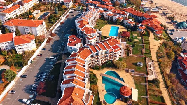 Aerial view of resort complex with swimming pools near the beach. Drone perspective of white buildings with orange tile rooftops, parking lot, and landscaped gardens.