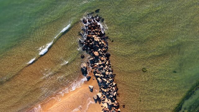 Stone jetty reaching into turquoise water from sandy shoreline. Overhead drone view of rock breakwater with sand patterns and gentle waves on both sides.