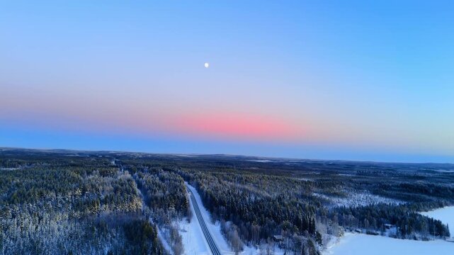 Road through boreal forest under pastel winter sky, Finnish Lapland. Aerial view of straight highway bisecting snowy taiga with moon and pink horizon band.