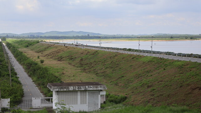 The image depicts a wide landscape featuring a sloped embankment running parallel to a water body, with a road lined with streetlights on the top edge of the embankment