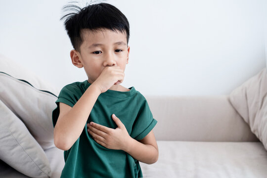 Sick Asian little boy coughing and holding his chest on a sofa. Child having respiratory illness, cold, flu, asthma symptoms, or bronchitis infection at home. Pediatric healthcare concept.