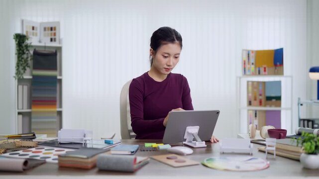 Asian woman interior designer furniture specialist using tablet computer reviewing design samples material swatches working at creative studio desk home office