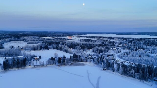 Frost covered village beside frozen lake under winter moon, Lapland. Aerial view of scattered houses, red barns and snowy fields framed by icy birch and spruce trees.