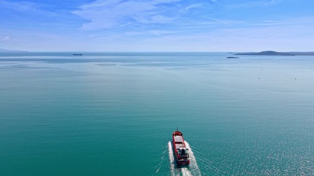 Cargo vessel heading toward open sea on calm waters. Rear aerial view of red freighter with white wake, sparkling surface and islands on the horizon.