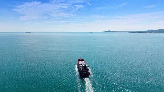Red cargo ship cruising through open sea near hilly coastline. Aerial view of lone vessel creating wake on glassy turquoise water with distant mountains.