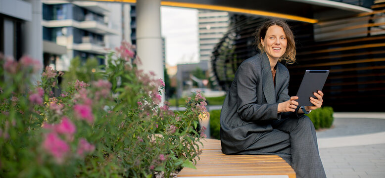 Woman uses tablet while sitting on bench in urban area with flowers around