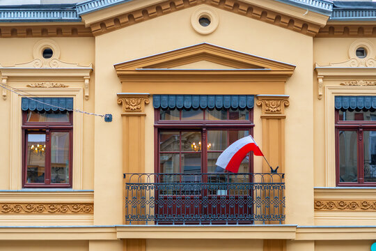 Historic yellow and orange building facade featuring ornate moldings, pilasters, and a wrought iron balcony railing with a waving red and white Polish flag, captured in daylight.