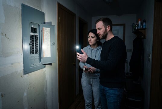 Couple inspecting an electrical breaker panel during a power outage at home, using smartphone and flashlight to troubleshoot household electricity issues in a dark residential hallway.