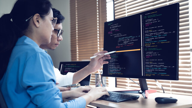 Two professionals focus intently on code displayed across dual monitors. One uses a stylus to point at highlighted lines of script