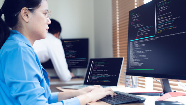 Woman types code at dual monitors in quiet office. Focus and concentration fill her calm expression. She works among colleagues on similar tasks. Perfect for tech, software, or developer projects