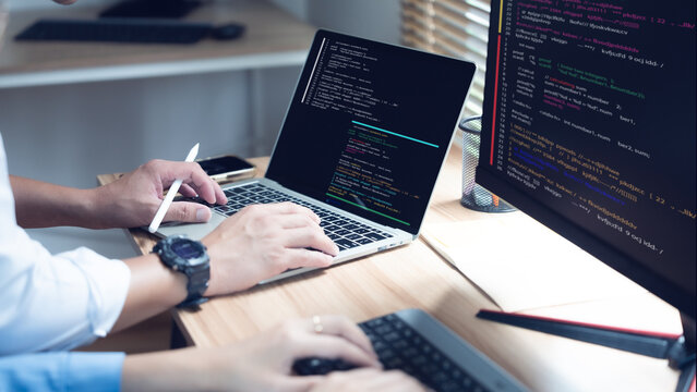 Coder types on laptop with keyboard and monitor nearby. Hands rest on keys while stylus hovers above screen. Code lines glow in darkened workspace. Fingers move purposefully as programmer focuses