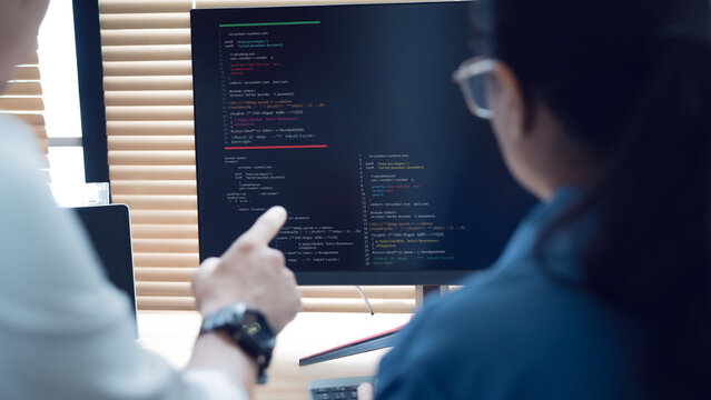 Two developers collaborate at a desk. One points at code on a large screen. Their focus shows teamwork and problem-solving. Soft lighting enhances the focused atmosphere. Ideal for tech, development
