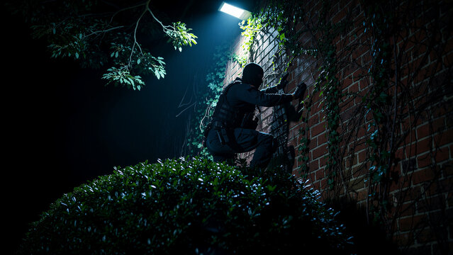Tactical intruder in balaclava scaling a brick wall at night under spotlight. Masked figure in gear climbs ivy-covered building exterior. Security breach concept for law enforcement or crime media.