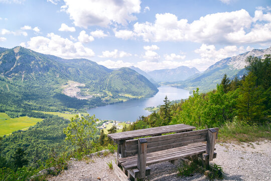 Panoramic view of Lake Grundlsee from the Lustiges Eck viewpoint in the Salzkammergut region of Styria, Austria. Wooden bench overlooking the alpine landscape with mountains, forest, and blue sky 