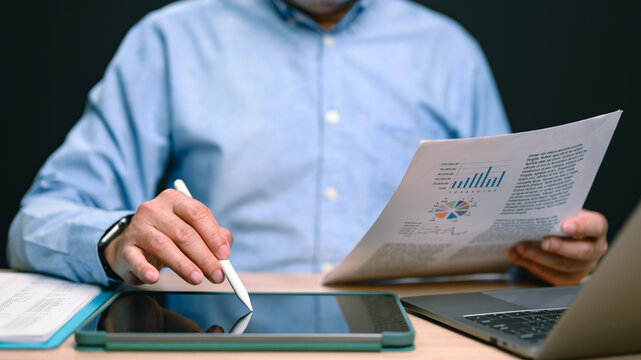 Professional male analyst sitting indoors reviews financial data reports holding paper charts employing stylus pen tablet screen near laptop computer inside dark office workspace evening work.