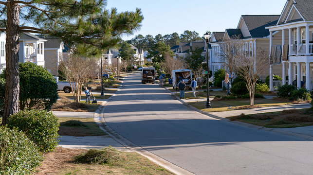 Wide-angle view of a suburban street in early spring with residents at various properties raking dead grass, pressure-washing driveways, airing out rugs over railings, and loading