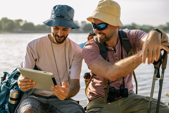 Gay male couple sitting under tree and looking at tablet during coastal hike