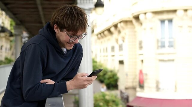 Side view of a smiling young man wearing glasses and a dark hoodie while looking at his smartphone. He stands outdoors in a city under a bridge structure during daytime.