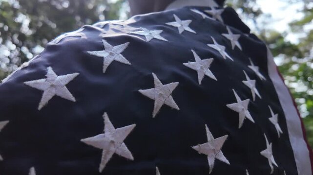 Close up side profile angle tracking the embroidered white stars on the blue field of an American flag being worn as a cape by a person standing outside.
