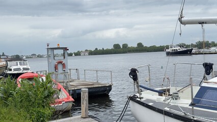 Gray sky hovers over calm water. Small boat rests near wooden dock. Metal pier extends into quiet lake. Greenery lines distant shoreline. Serene, peaceful scene ideal for travel or nature themes © KARINA