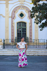Obraz premium A young, beautiful woman with long dark hair tied back in a ponytail, wearing a white top and a long floral-print skirt, poses like a model next to an old building in the centre of Seville, Spain.