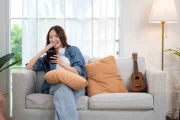 Obraz premium Young Asian woman relaxing on a sofa with a smartphone and headphones beside a ukulele in a bright living room