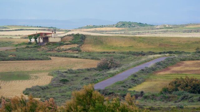 Old abandoned farmhouse with tiled roof sits above a winding country road, patchwork of cultivated fields and dry grassland spreads across rolling terrain toward hazy hills