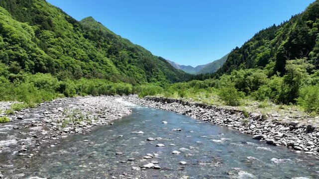 Clear river flows through rocky valley. Lush green mountains rise on both sides. Bright blue sky frames the natural scene. Rushing water suggests energy and life