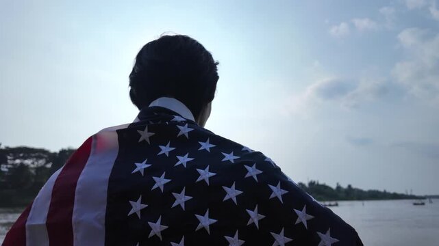 Medium shot from behind showing the silhouette of a man draped in the United States flag like a cape, standing still on the riverbank while looking out over the calm water under a bright sunny sky.
