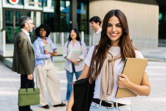 Portrait of young adult university female student smiling at camera holding notebook on campus. Young woman standing outdoors before class in a modern educational setting. Student lifestyle concept.