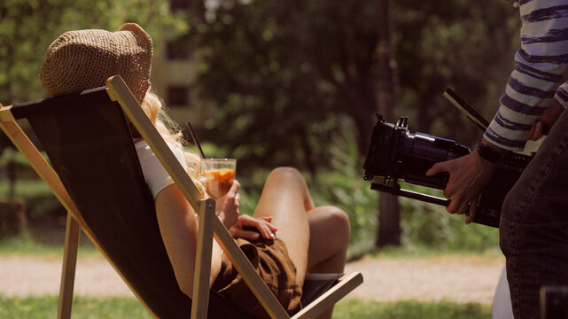 Woman sitting on a lounge chair in an outdoor cafe. Holding an iced coffee with a straw. Man with a film camera taking photos