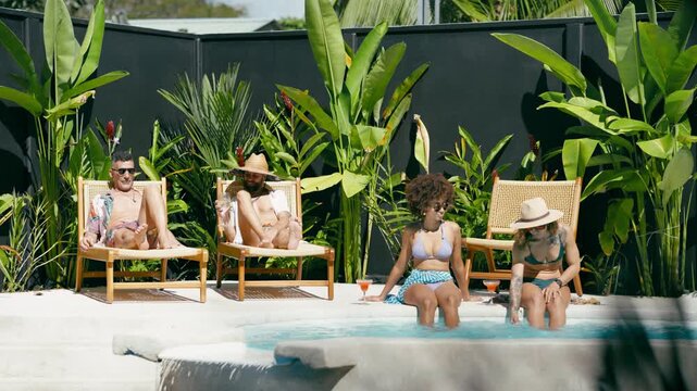 Group of four diverse friends sunbathing and relaxing by a luxury swimming pool. Two men tanning on lounge chairs and two women in bikinis with cocktails